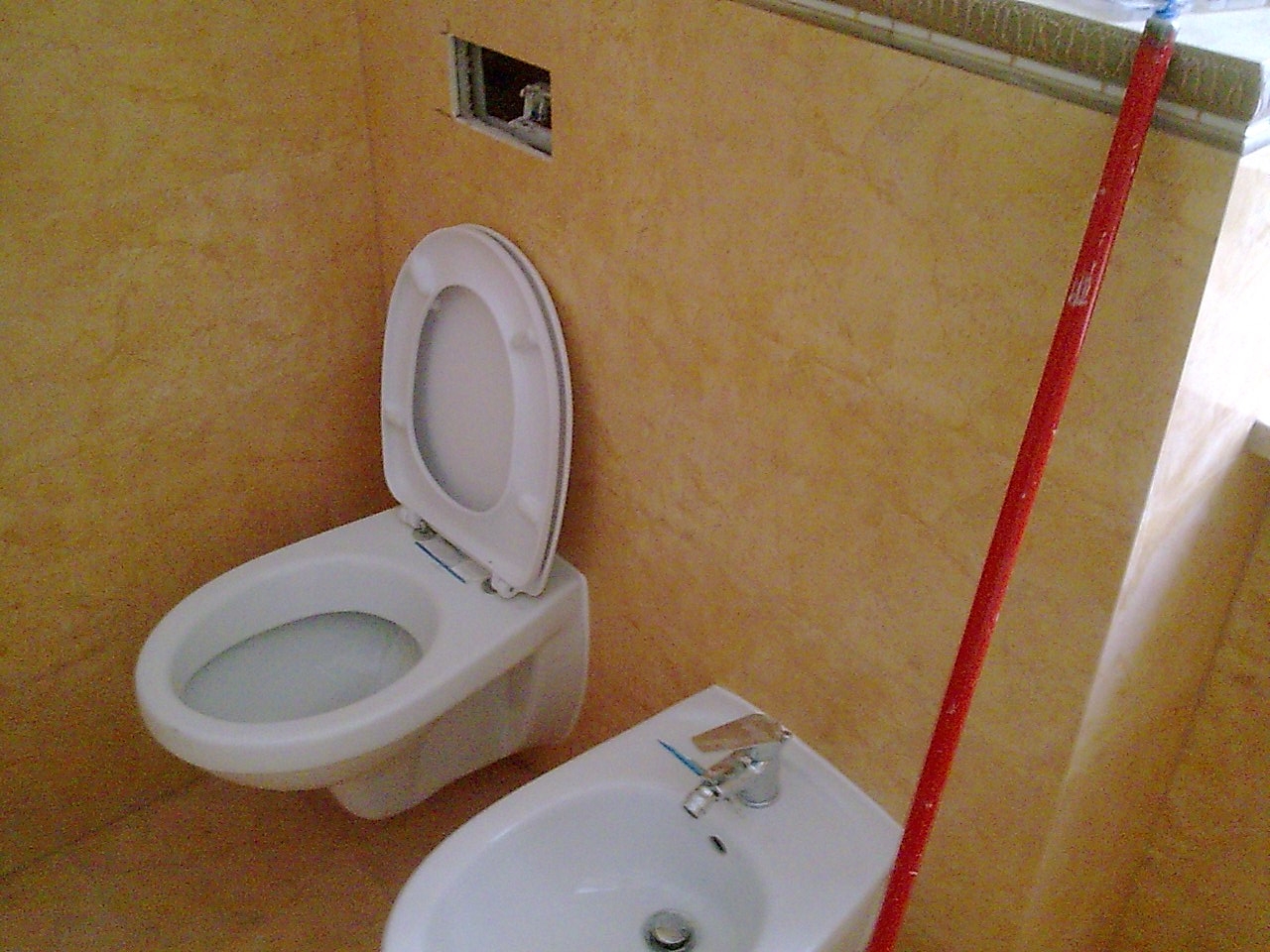 Zamontowana biała toaleta podwieszana z otwartą deską i bidet w łazience wykończonej w odcieniach beżu; widoczny fragment czerwonego kija oparty o ścianę.